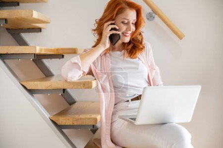 Téléchargez les photos : Joyeux et belle jeune femme parlant au téléphone et travaillant sur un ordinateur portable sur ses genoux tout en étant assis dans les escaliers à la maison - en image libre de droit