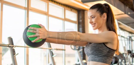 Young fit woman doing bent knee exercise with ball in gym. Beautiful smiling woman doing exercise with medicine ball wearing sportswear and ponytail in gym.