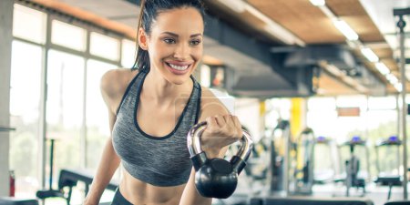 Photo for Smiling young woman working out with kettle bell in gym. Beautiful fitness girl in sportswear working out with kettle bell in the morning. - Royalty Free Image