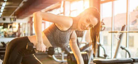 Téléchargez les photos : Jeune femme en forme en tenue de sport travaillant avec des poids sur banc de fitness dans la salle de gym. Fille souriante attrayante travaillant devant la lumière du soleil le matin. - en image libre de droit