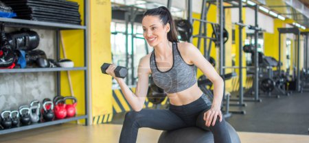 Photo for Beautiful young woman lifting weights in gym and sitting on fitness ball. Young attractive woman working out with weights at gym. - Royalty Free Image
