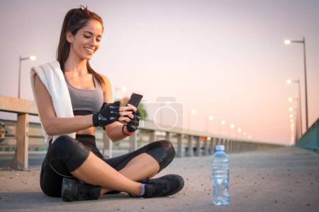 Téléchargez les photos : Belle sportive souriante prenant une pause après l'entraînement et en utilisant le téléphone à l'extérieur. - en image libre de droit