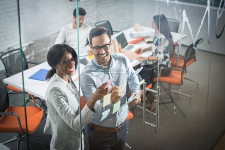 Foto de Dos colegas de negocios haciendo una lluvia de ideas juntos en una pared de vidrio con notas adhesivas mientras están de pie en una oficina moderna - Imagen libre de derechos