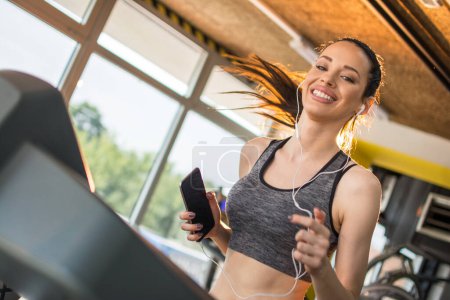 Photo for Attractive young sportswoman with earphones and smartphone running on a treadmill at gym - Royalty Free Image