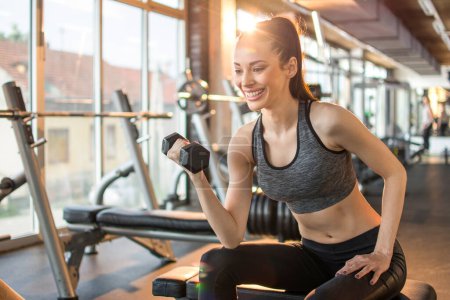 Téléchargez les photos : Femme sportive active flexion des muscles avec haltère assis sur un banc de musculation dans la salle de gym - en image libre de droit