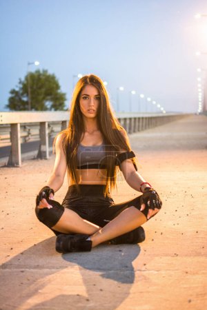 Téléchargez les photos : Belle jeune sportive assise sur le trottoir et relaxante après avoir couru sur le pont le soir. - en image libre de droit