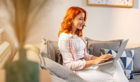 Photo for Young ginger smiling woman sitting on sofa and typing on laptop. Working from home. - Royalty Free Image