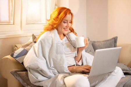 Foto de Feliz joven pelirroja bebiendo café cubierto con manta mientras trabaja en el ordenador portátil y sentado en el sofá en casa. - Imagen libre de derechos