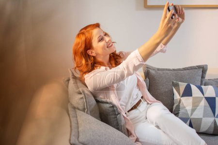 Téléchargez les photos : Heureuse jeune femme rousse se détendre, prendre selfie et assis sur le canapé à la maison. - en image libre de droit