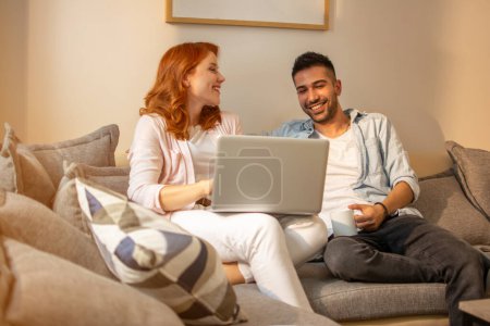 Téléchargez les photos : Mignon couple heureux en utilisant un ordinateur portable tout en étant assis sur le canapé à la maison. - en image libre de droit