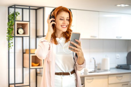 Photo for Young, ginger, beautiful and happy girl in bright outfit enjoying to the music with headphones on her head, while using her smartphone and standing at home - Royalty Free Image