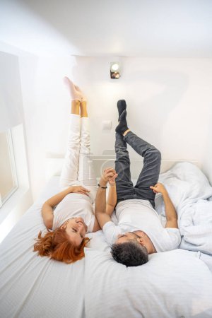 Foto de Joven feliz hombre y mujer acostados en la cama en casa levantando las piernas y tomados de la mano. - Imagen libre de derechos