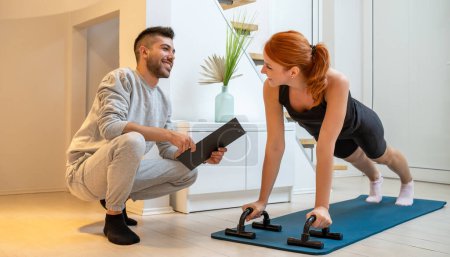 Photo for Happy beautiful ginger woman doing home workout, push up exercise with assistance of her personal trainer at home in living room. - Royalty Free Image