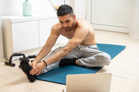 Foto de Joven hombre guapo haciendo ejercicio de estiramiento mientras ve entrenamiento deportivo en línea en el ordenador portátil en casa - Imagen libre de derechos