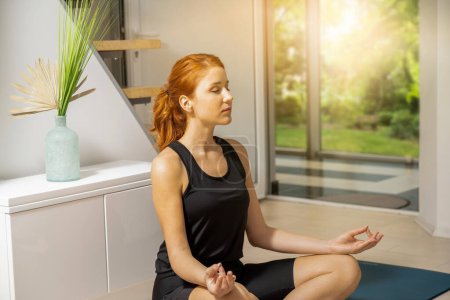Photo for Attractive young ginger woman with closed eyes exercising and sitting in yoga lotus position while resting at home. - Royalty Free Image