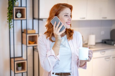 Photo for Portrait of young, ginger, beautiful and happy girl drinking tea while talking on the phone and standing in kitchen at home in the morning - Royalty Free Image