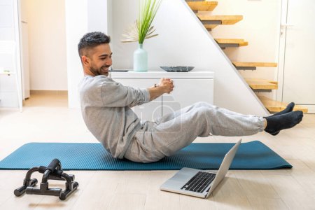 Feliz joven guapo haciendo ejercicio de giro ruso en la alfombra de ejercicio y viendo clases de deporte a través de la computadora portátil en casa.