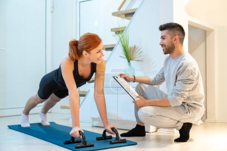 Photo for Smiling beautiful ginger woman doing home workout, push up exercise with assistance of her personal trainer at home in living room. - Royalty Free Image