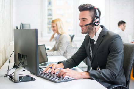 Foto de Joven operador de soporte al cliente masculino guapo en ropa de negocios con auriculares hablando con el cliente mientras trabaja en la computadora en su lugar de trabajo - Imagen libre de derechos