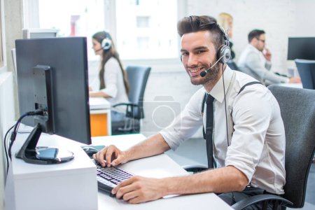 Foto de Operador de soporte al cliente joven guapo con auriculares manos libres trabajando con computadora en el centro de llamadas - Imagen libre de derechos