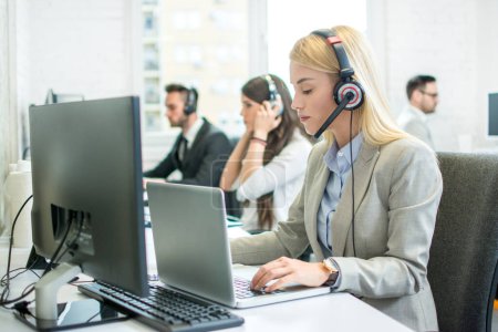 Foto de Hermosa mujer joven operador de atención al cliente con auriculares que trabajan en el ordenador portátil en el centro de llamadas - Imagen libre de derechos