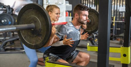Photo for Side view of muscular handsome man lifting heavy barbell with assistance of young attractive fit woman at the gym. - Royalty Free Image