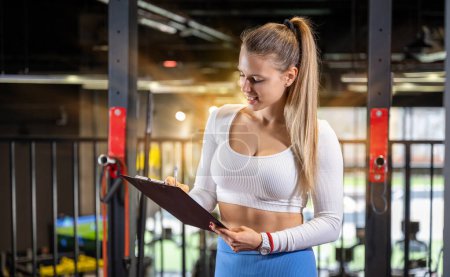 Photo for Young fitness instructor making schedule for a client on the clipboard at the gym. Attractive blonde trainer writing report for a client. - Royalty Free Image