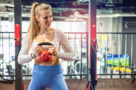 Téléchargez les photos : Portrait de jeune femme blonde faisant de l'exercice avec kettlebell à la salle de gym. Femme sportive travaillant sur le port de vêtements de sport. - en image libre de droit