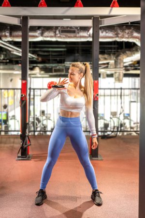 Foto de Mujer rubia joven haciendo ejercicio con kettlebell en el gimnasio. Mujer deportiva haciendo ejercicio usando ropa deportiva. - Imagen libre de derechos