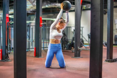 Téléchargez les photos : Attrayant jeune femme debout sur ses genoux faire de l'exercice avec balle de fitness à la salle de gym. - en image libre de droit