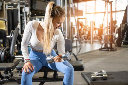 Téléchargez les photos : Jeune femme de fitness soulevant du poids à la salle de gym. Femme blonde sportive assise sur un banc de musculation et soulevant des poids au gymnase. - en image libre de droit
