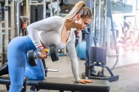Foto de Atractiva mujer en forma levantando pesas en el banco del gimnasio. Mujer deportiva rubia haciendo ejercicio con pesas. - Imagen libre de derechos