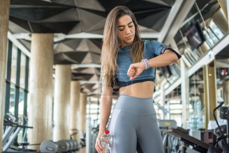 Téléchargez les photos : Jeune sportive vérifiant son activité physique et brûlant des calories sur smartwatch après l'entraînement dans la salle de gym - en image libre de droit
