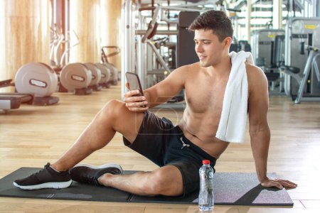 Handsome shirtless young guy resting after training and using phone at gym
