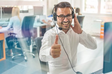 Photo for Portrait of excited man sitting at his workplace showing thumbs up sign gesture, wearing headphones posing to camera. Hot line support operator at call center - Royalty Free Image