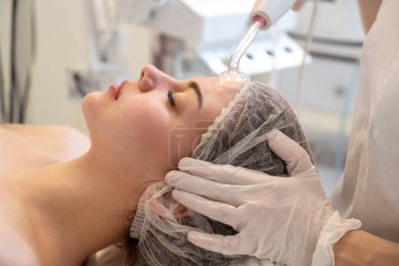 Foto de Esteticista aplicando tratamiento de masaje facial eléctrico Darsonval con herramienta especial sobre la frente de las pacientes femeninas en el salón de belleza - Imagen libre de derechos