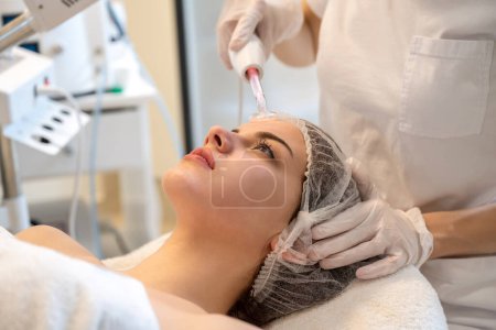 Téléchargez les photos : Esthéticienne appliquant un traitement électrique de massage facial Darsonval avec un outil spécial sur le visage des patients dans un salon de beauté - en image libre de droit