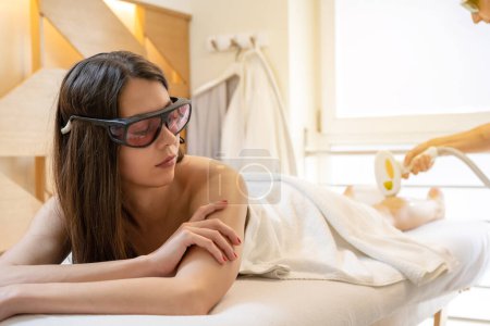 Téléchargez les photos : Jeune femme avec des lunettes de protection obtenir l'épilation au laser sur ses jambes dans le salon de beauté - en image libre de droit