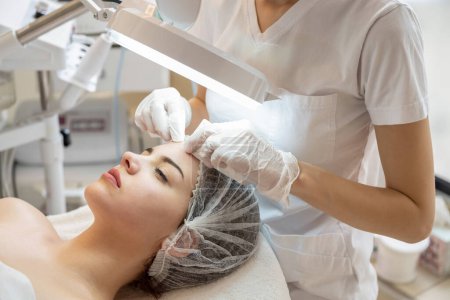 Téléchargez les photos : Cosmetologue serrant l'acné sur le front des patientes avec des lingettes dans le salon de beauté - en image libre de droit