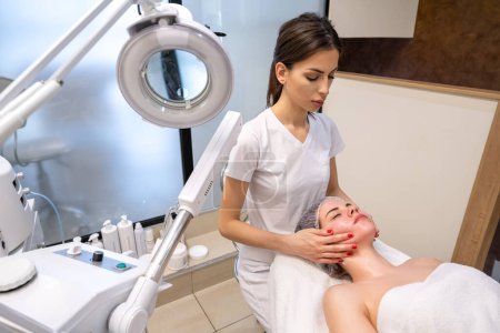 Téléchargez les photos : Thérapeute appliquant un massage curatif des joues faciales à la jeune femme dans un salon de beauté. - en image libre de droit