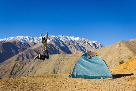Téléchargez les photos : Excited young man having a blast jumping next to igloo camping tent with splendid views of Andes snowed mountain range under intense blue sky in Elqui Valley, Chile. Adventure, hiking concept - en image libre de droit
