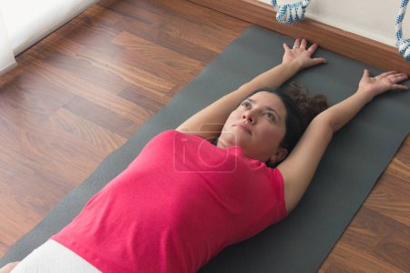Foto de Young female yogi lying down on mat stretching back with arms above head. Yoga instructor on pink t-shirt warming up for practice. Healthy lifestyle, exercise concept - Imagen libre de derechos