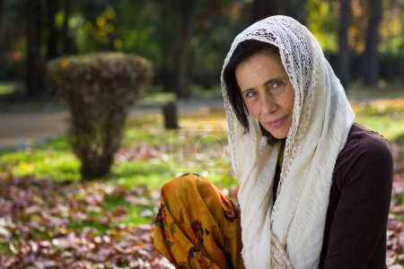 Foto de Precioso retrato de mujer madura con chal blanco sobre la cabeza sentado en hojas marrones en el parque. Edad media hermosa dama con mirada musulmana al aire libre - Imagen libre de derechos