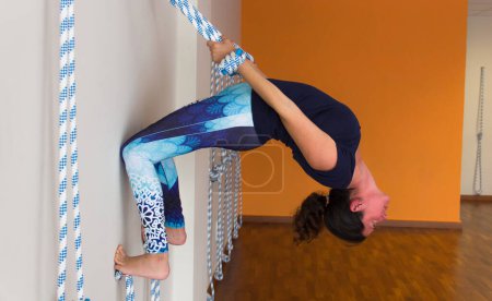Téléchargez les photos : Young yogi grabbed to ropes and hanging from white wall doing back bending in yoga center. Advanced yoga teacher on upside down challenging posture in empty studio - en image libre de droit