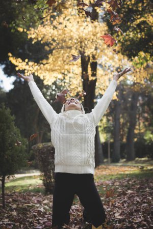 Foto de Euphoric woman stands on her knees with open arms looking up while brown leaves float in the air during fall season in the park. Autumn season fashion sales concept - Imagen libre de derechos