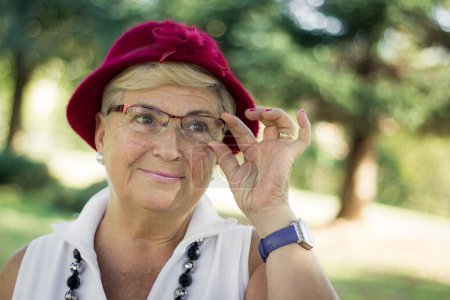 Photo for Smiling senior woman with red hat holding eyeglasses outdoors. Pretty older blonde lady on sunny morning in the park. Young looking retired person concept - Royalty Free Image