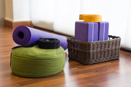 Téléchargez les photos : Various yoga props on studio wood floor. Set of blocks in wicker basket, belts, mat and green cushion in yoga center. Wellness activity concept - en image libre de droit