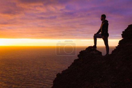 Téléchargez les photos : Hiker silhouette standing alone on cliff edge rock staring at sunset in Gran Canaria, Spain. Fearless man enjoys splendid twilight by ocean in Canary Islands. Visionary, adventure, challenge concepts - en image libre de droit