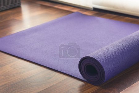 Foto de Purple yoga mat half unrolled on wooden floor by white curtain window in yoga studio. Practice exercise, healthy lifestyle concept - Imagen libre de derechos