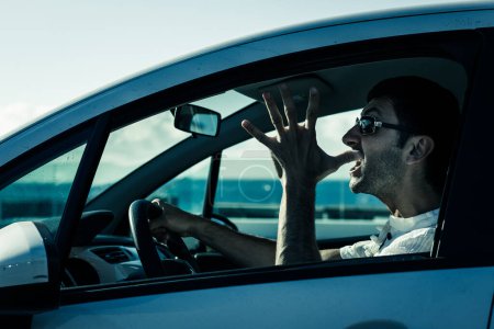 Photo for Furious driver gesticulates with open hand on white car. Desperate young man with sunglasses driving with anger. Upset, aggressive, impatient, bad manners concepts. Cold blue effect on sunny day - Royalty Free Image